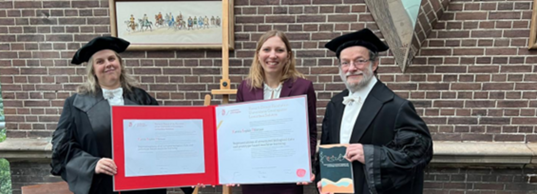 Three people, two of whom are wearing academic gowns, are standing by a brick wall, holding large picture frames in their hands