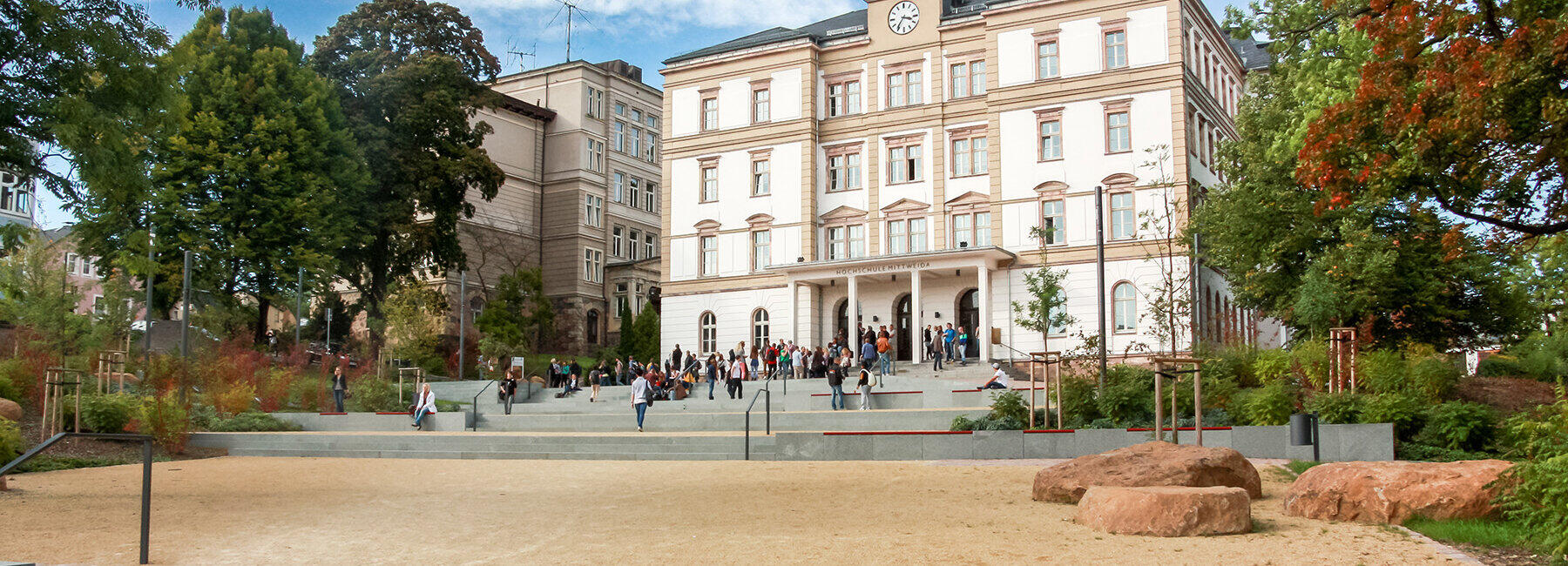 Großes historisches Gebäude mit mehreren Stockwerken, davor ein offener Platz mit Bäumen und Bänken