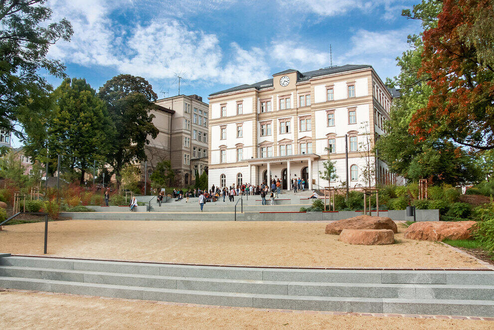 Großes historisches Gebäude mit mehreren Stockwerken, davor ein offener Platz mit Bäumen und Bänken