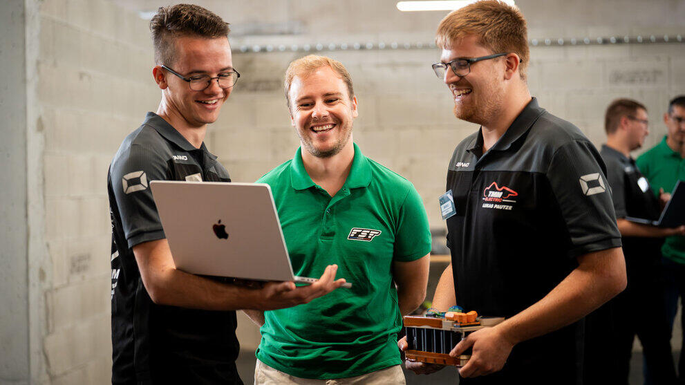 Drei männliche Personen schauen lachend auf einen geöffeneten Laptop