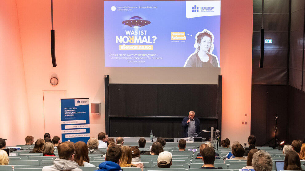 Blick von hinten über die Köpfe der Besucher:innen hinweg in den Hörsaal. Hinter dem Pult spricht eine männliche Person mit Mikrofon in der Hand zum Publikum. Links steht ein blaues Rollup u.a. mit dem Text „Institut für Kompetenz, Kommunikation und Sprachen“. Das Beamerbild an der Wand zeigt die Begrüßungsfolie u.a. mit dem Text „Was ist normal? Ringvorlesung“ und dem Titel der ersten Vorlesung: „Das ist so ein warmes Heimatgefühl“ – Sozialpsychologische Perspektiven auf die Suche nach Normalität“. Daneben ist die Referentin Henriette Rodemerk abgebildet.