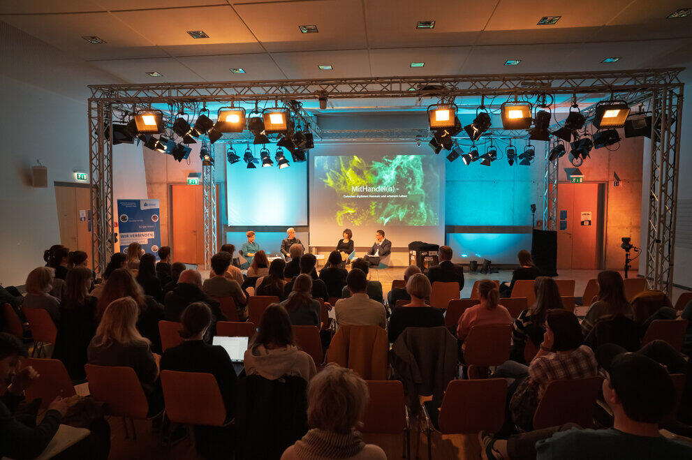 Publikum sitzt in einem Saal vor einer Bühne mit vier Personen in Podiumsdiskussion und einer großen Leinwand mit grünem Bildhintergrund