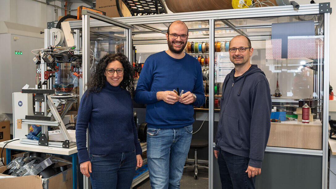 Eine weibliche Person links und zwei männliche Personen stehen lächelnd vor einem gläsernen Laborabteil, in dem sich unter anderem zahlreiche Rollen mit farbigem Filament für den 3D-Druck befinden.