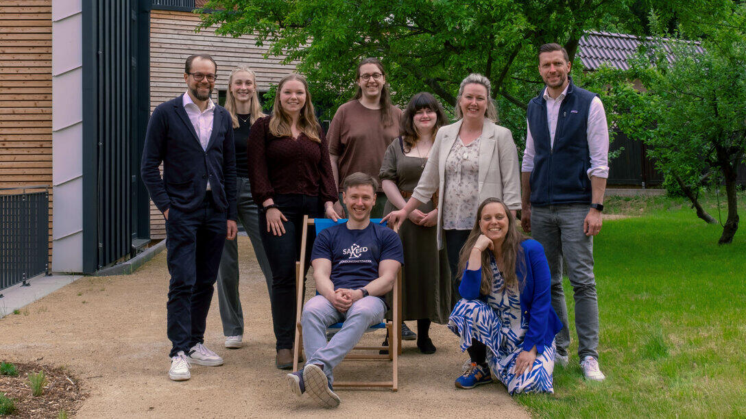 Gruppenbild mit neun Personen vor einem Gebäude und einer Wiese mit Bäumen.
