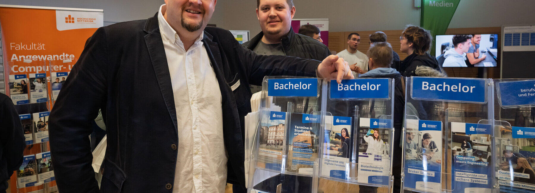 Eine ältere und eine jüngere männliche Person stehen lächelnd an einem Ständer mit vielen Studiengangsflyern