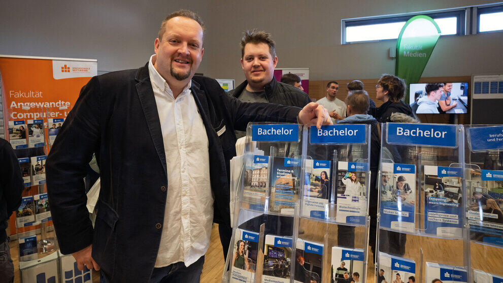 Eine ältere und eine jüngere männliche Person stehen lächelnd an einem Ständer mit vielen Studiengangsflyern.