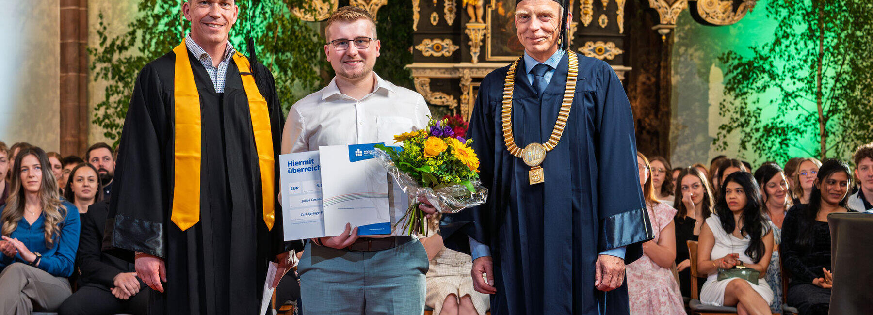 Three men standing in the chancel of a church. The younger man in the middle is holding a bouquet of flowers and a certificate. The men on the left and right are wearing academic gowns and caps; the man on the right is also wearing a gold chain of office.