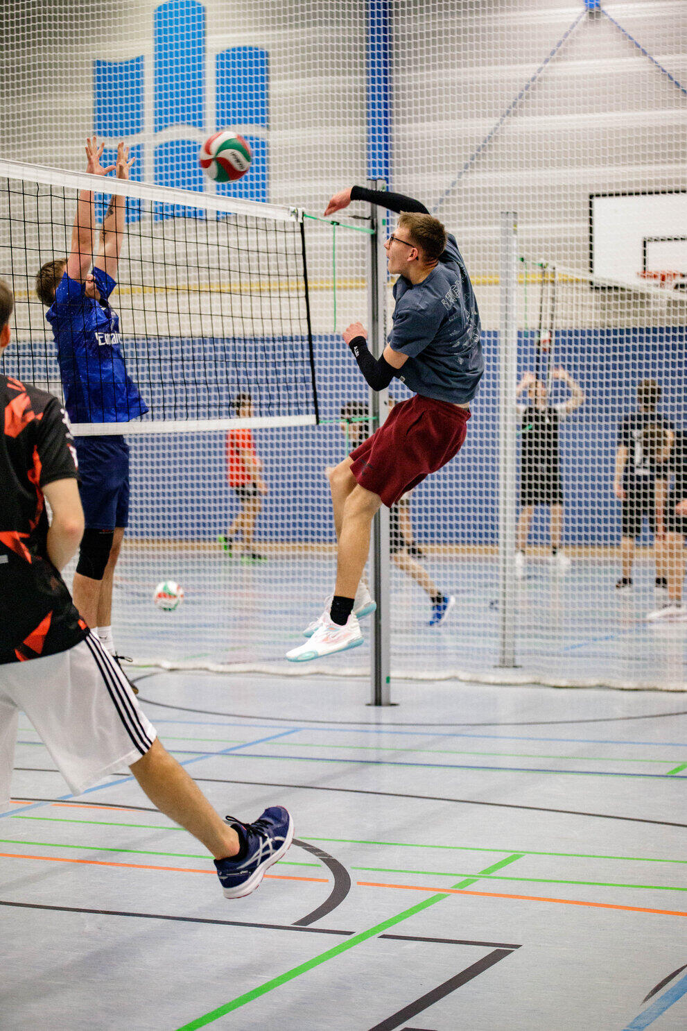Ein Mann schmettert den Volleyball übers Netz, sein Gegenspieler versucht zu blocken. Im Hintergrund hängt das Logo der Hochschule Mittweida an der Sporthallenwand.
