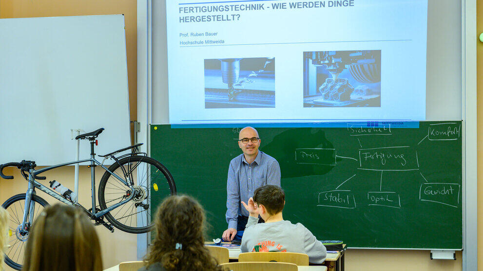 Blick von hinten über Schüler:innen in einem Klassenraum hinweg auf eine männliche Person vor einer Tafel, neben ihr ein Fahrrad auf einem Ständer , hinter ihr eine projizierte Folie mit der Überschrift "Fertigungstechnik".