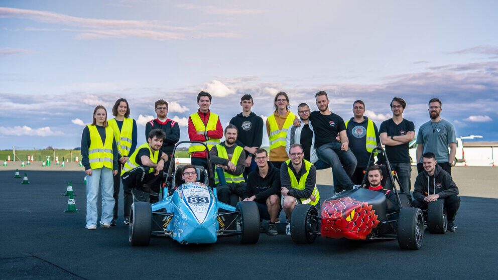 A group photograph showing 18 young people, some wearing yellow high-visibility vests, alongside a blue and a red open-top racing car on a tarmac surface. Two of the people are sitting in the racing cars, five are kneeling to the left and right of them, and the others are standing behind them. Red and white cones are set up in the background of the picture.