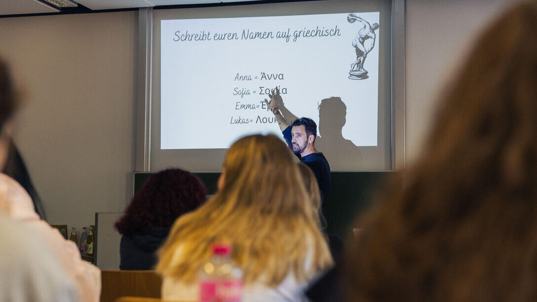 Blick durch die Zuhörenden auf einen Dozenten. Er zeigt auf eine projizierte Folie mit der Überschrift "Schreibe deinen Namen auf Griechisch" darunter die Namen Anna, Sofia, Emma und Lukas jeweils in lateinischen und griechischen Buchstaben.