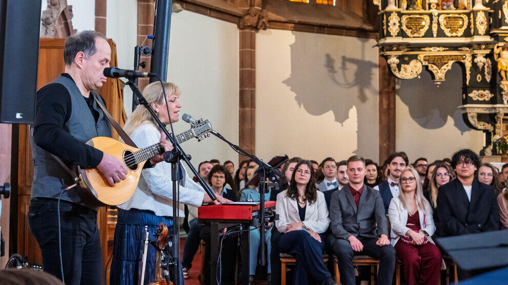 A musical duo is pictured in the foreground, to the side. Behind them, graduates are sitting on chairs.