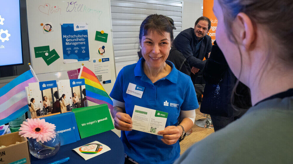 Lachende weibliche Person vor "International-Office" Rollup hält ein "I love Erasmus+"-Kärtchen in den Händen und unterhält sich mit zwei Personen im Bildvordergrund