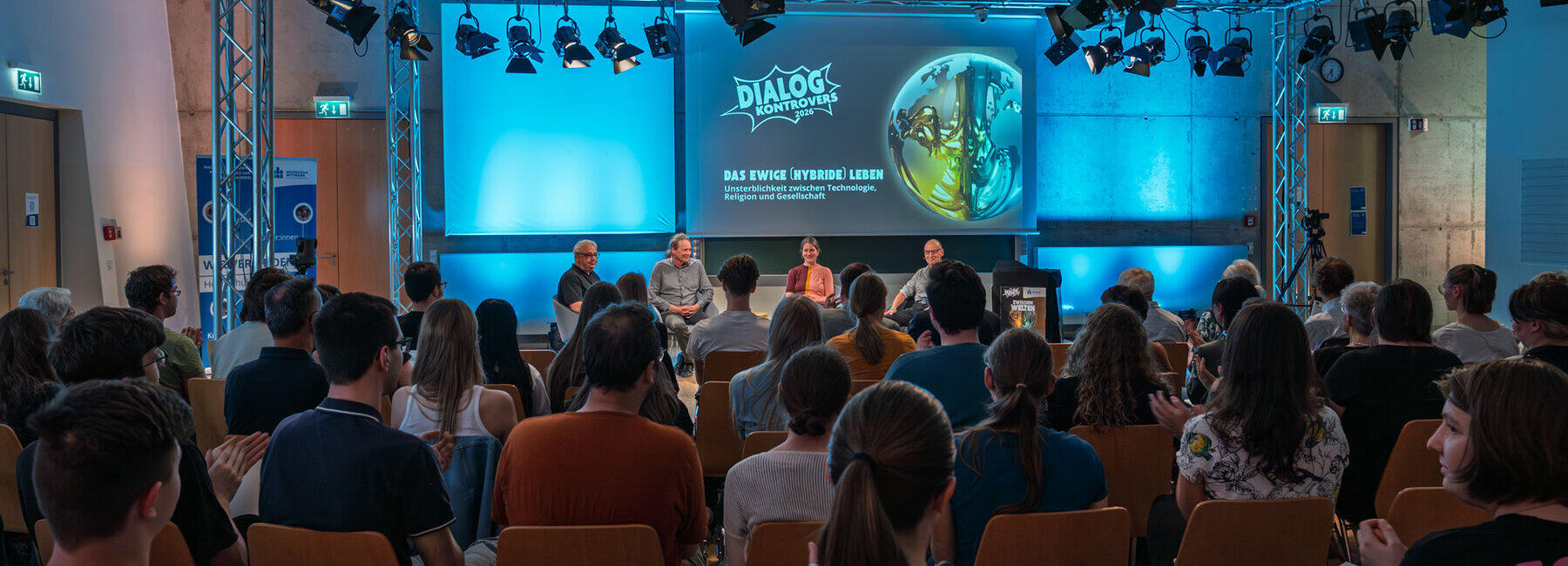 In einem großen Raum fällt der Blick über die Köpfe der Zuschauer:innen hinweg auf das mit vier Pesonen bestzte Podium unter an Traversen hängenden Scheinwerferen. Auf der Leinwand hinter ihnen ist das "Dialog Kontrovers"-Motiv und das Thema "Das ewige hybride Leben" projiziert.