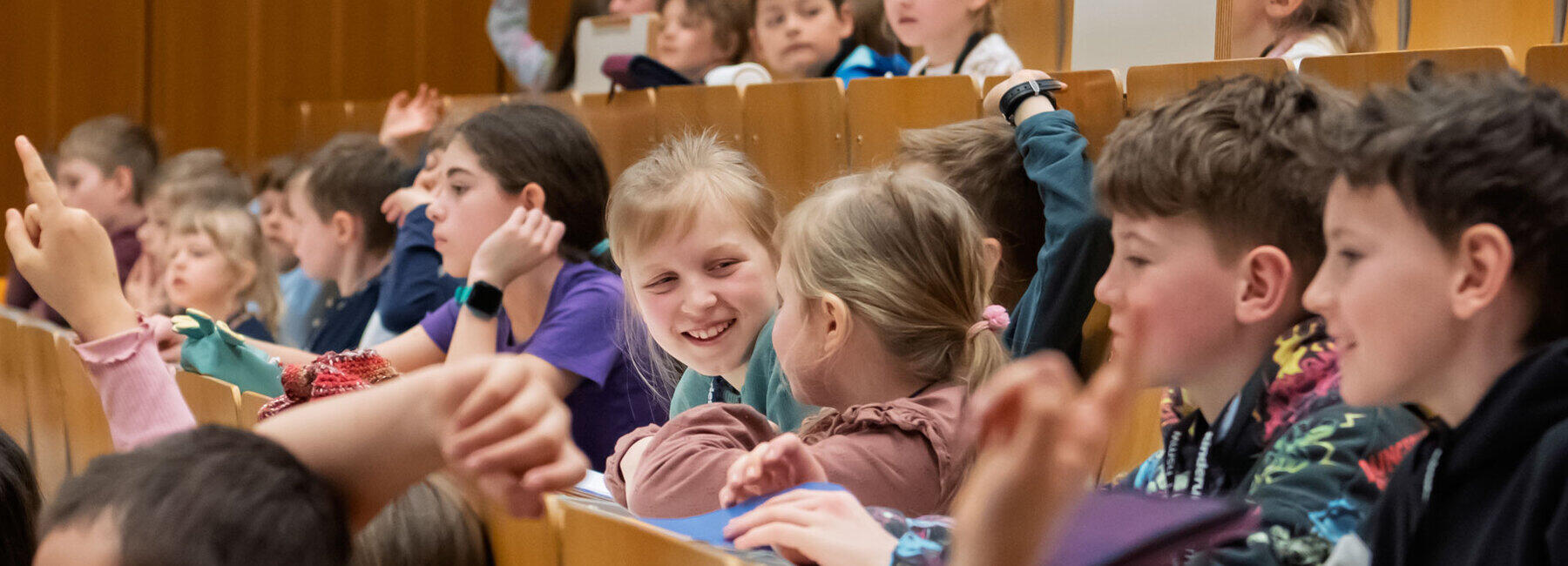 Gruppe von Kindern in den ansteigenden Bankreihen des Hörsaals. Einige melden sich mit gestreckten Armen, während andere miteinander sprechen und lachen.