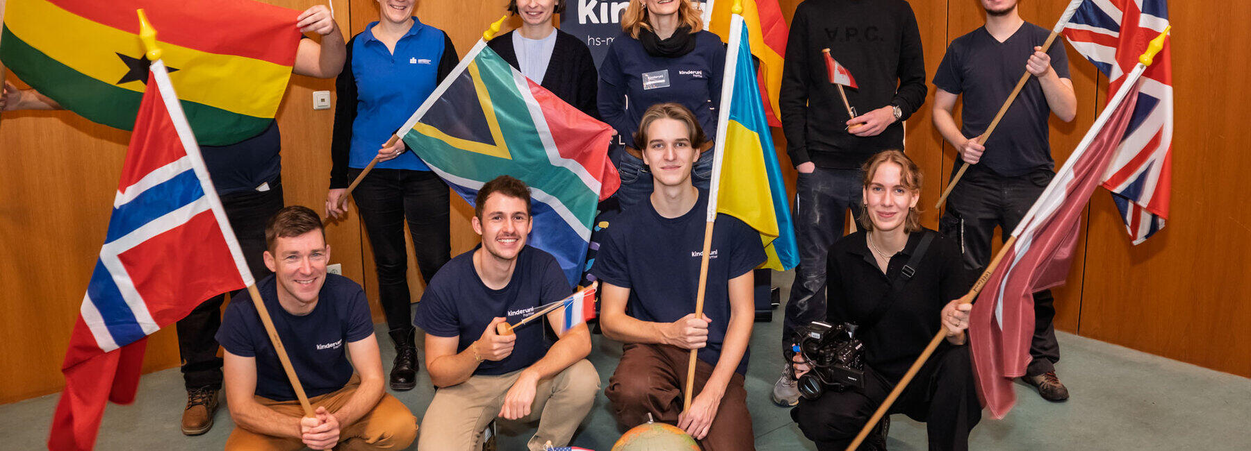 Zehnhn Personen stehen und knien im Hörsaal, einige halten verschiedene Nationalflaggen hoch und lächeln. Hinter der Gruppe ist ein Kinderuni-Rollup.