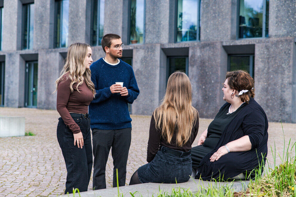Vier Studierende stehen und sitzen vor einem modernen Gebäude und unterhalten sich.