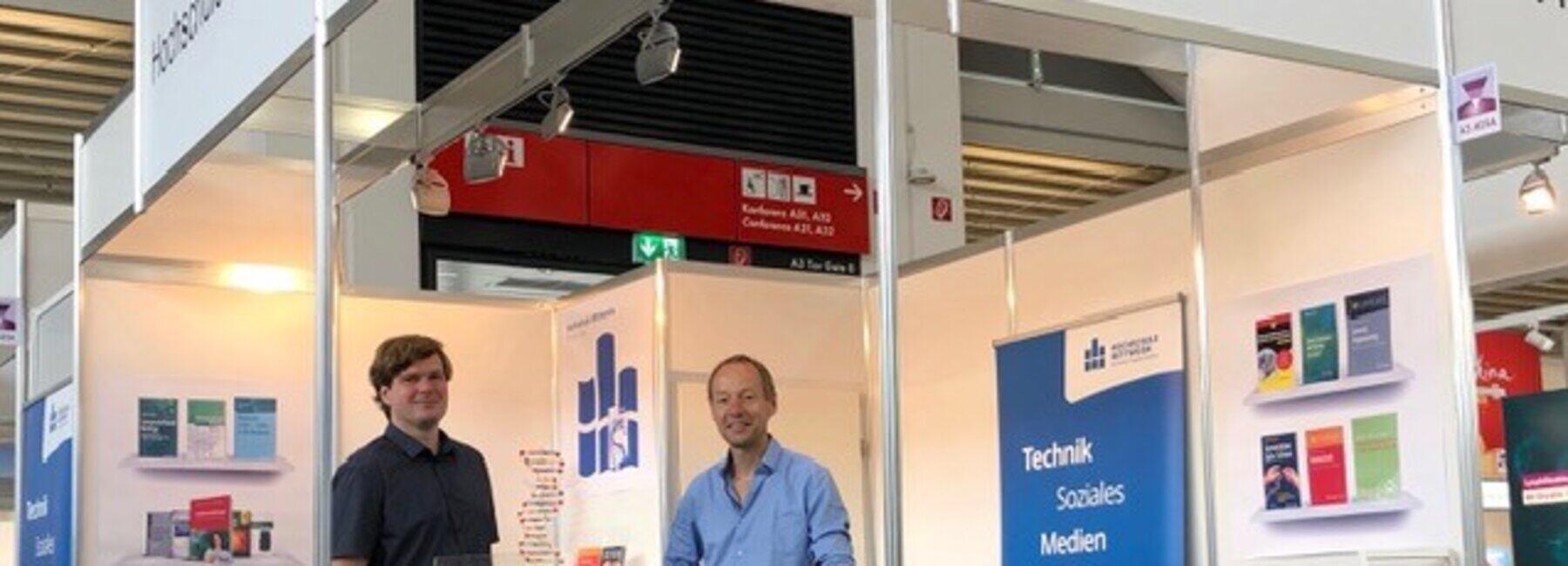 zwei männliche Personen stehen an einer Theke an einem Eck-Messestand mit Schriftbanner Hochschule Mittweida und Postern an den Wänden