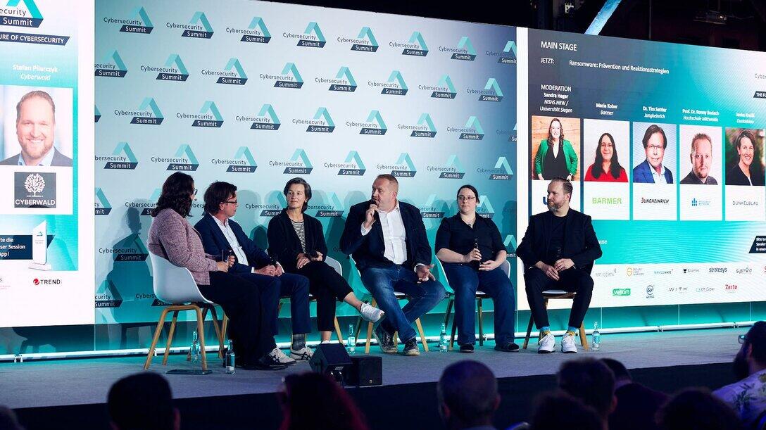 Blick auf ein Podium, 6 Personen sitzen auf Stühlen und diskutierenn
