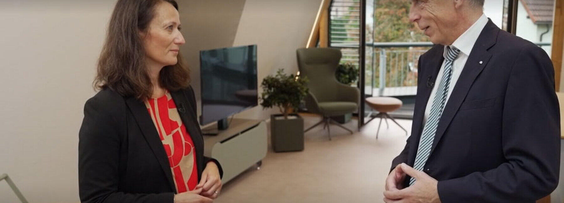 Eine weibliche und eine Person stehen sich im Gespräch in einem Raum mit einer großen Fensterfläche im Hintergrund gegenüber. in der oberen linken Bildecke ist das Sender-Logo "SR" zu sehen.