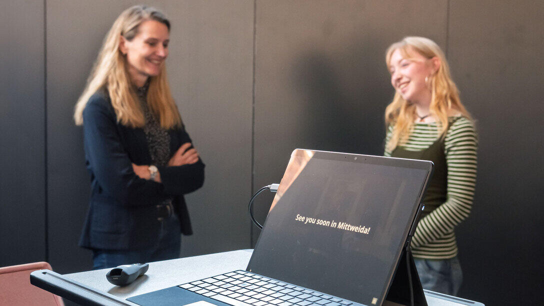 An open laptop on a desk. The screen displays the sentence: ‘See you soon in Mittweida’. In the background are two smiling women.