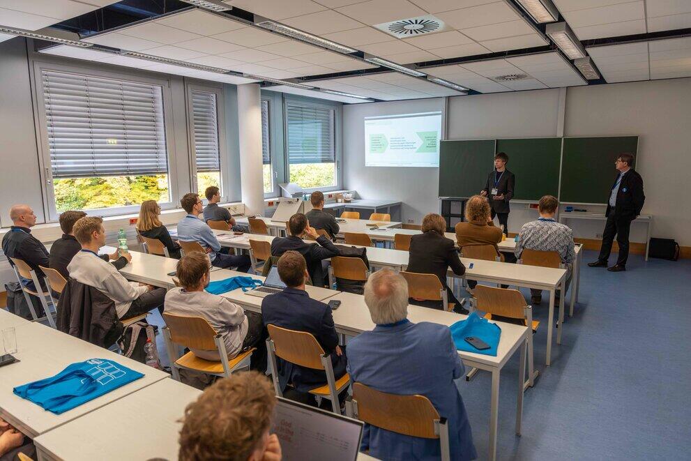Blick in einen gefüllten Seminarraum mit zwei vortragenden Personen an der Tafelseite