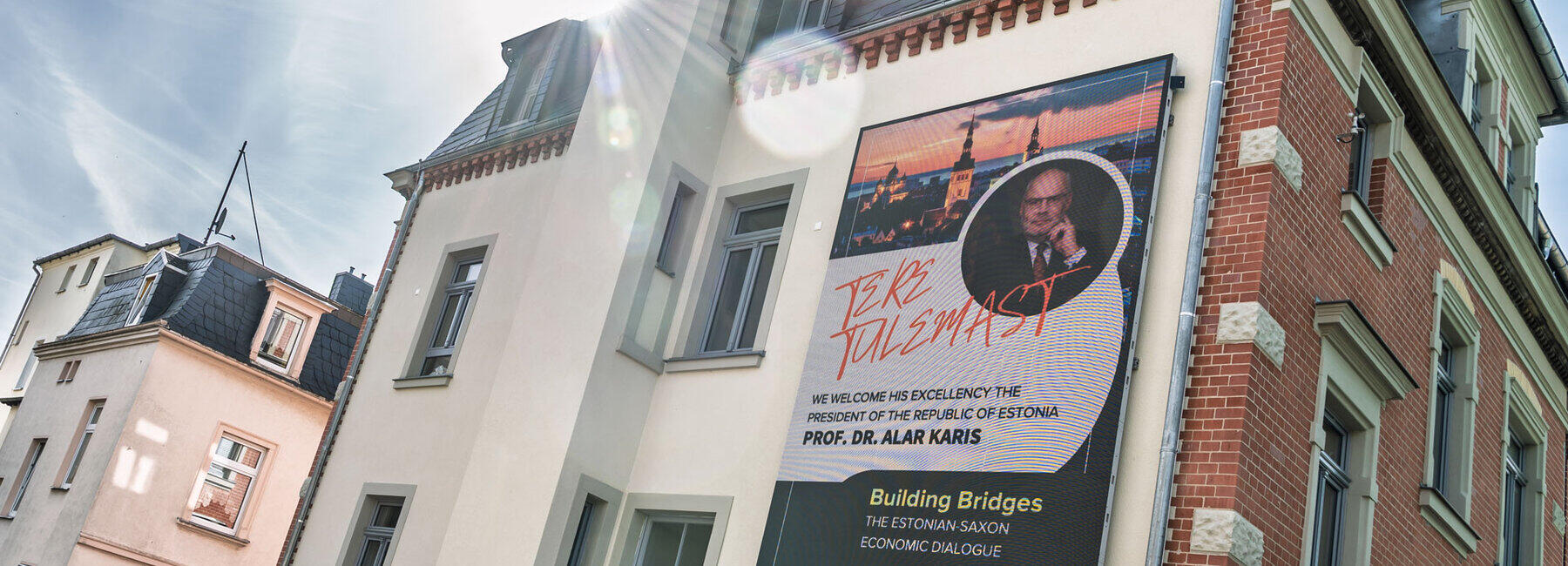 A building façade featuring a wall-sized LED display: it shows a photograph by Alar Karis and, amongst other things, the text ‘Building Bridges’. In the photograph, taken against the sun, the lenses of the camera are reflected.