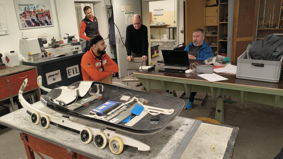 Vier männliche Personen stehe und sitzen in einer Werkstatt an Laptop und Rennschlitten