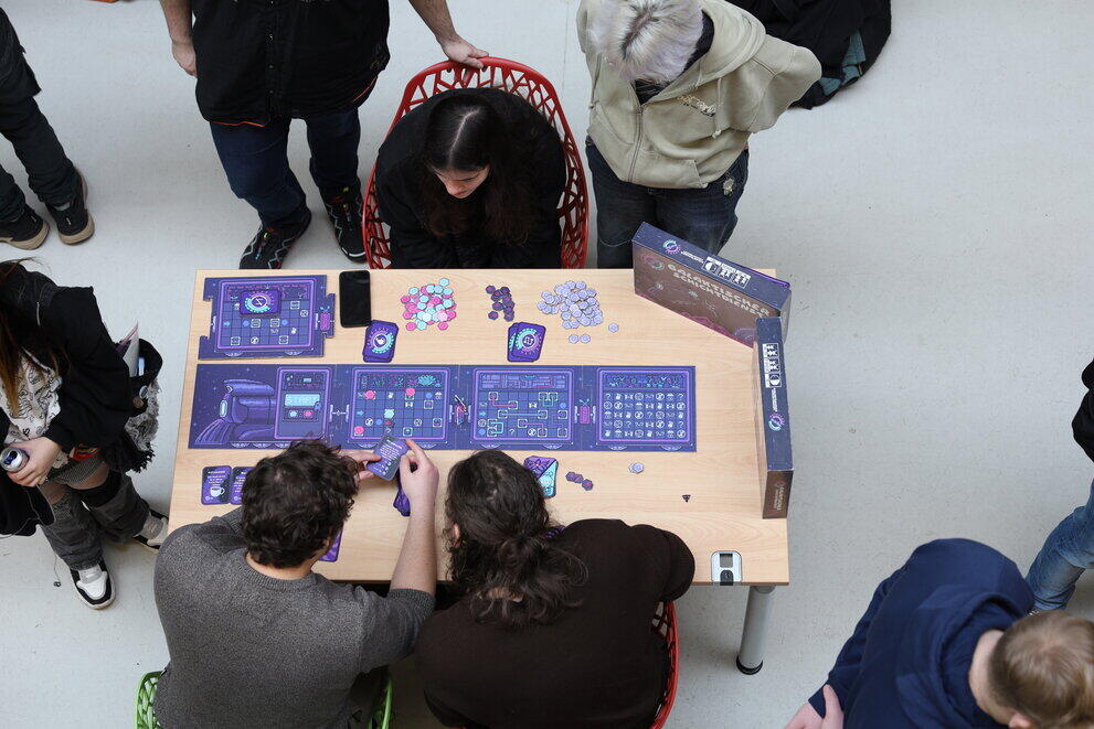 Viele Studierende stehen an einem Tisch und spielen ein Brettspiel.