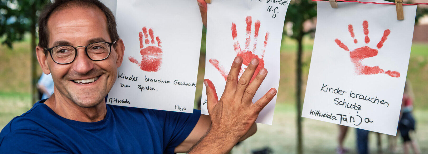 A man wearing glasses and a blue T-shirt stands in front of a washing line, smiling cheerfully, to which sheets of paper bearing handprints in red paint are attached with clothes pegs. Beneath the handprints are handwritten comments such as ‘Power to the children’ and ‘Children need protection’.