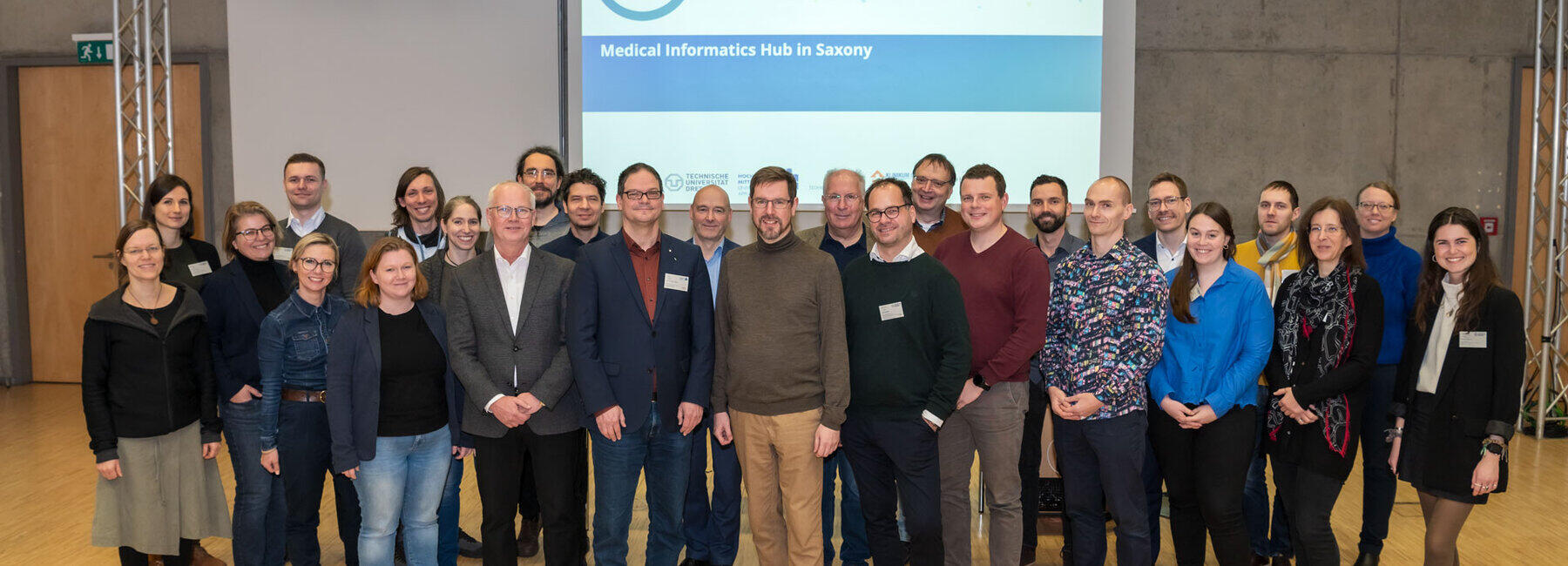 Gruppenaufnahme mit 26 Personen. In Hintergrund ist eine Folie projiziert mit dem MiHUBx-Logo und dem Schriftzug „Medical Informatics Hub in Saxony“.