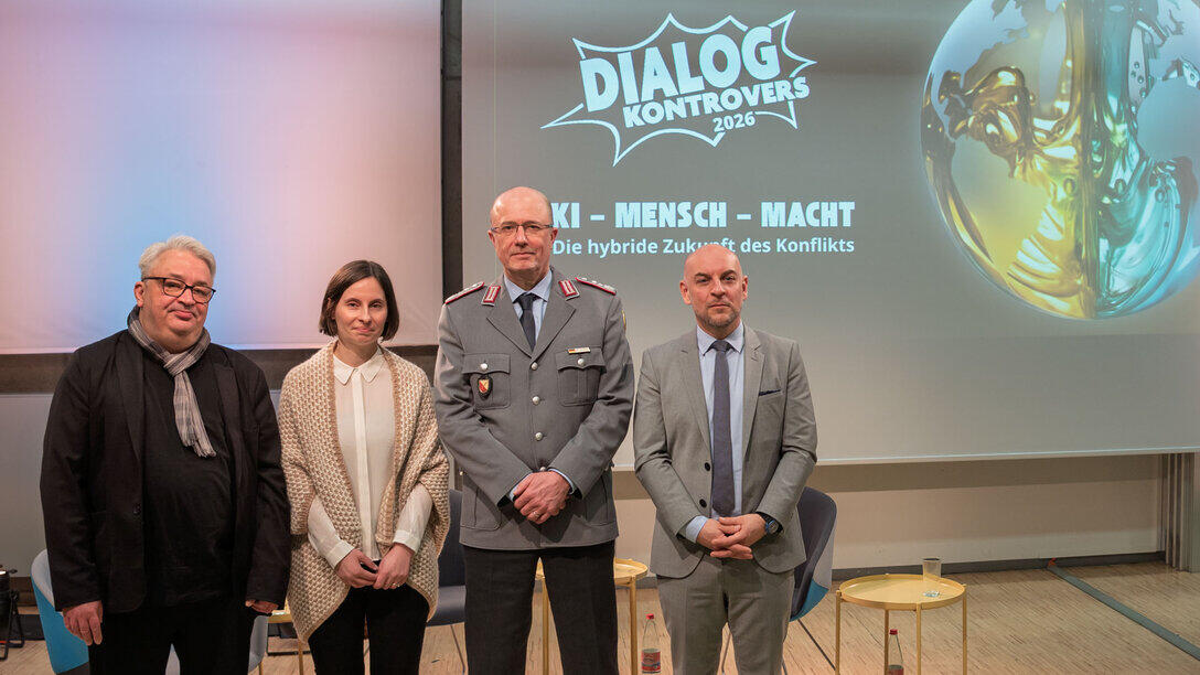 Four people, including one woman, are standing side by side for a group photo. On the screen in the background, the Controversial Dialogue (Dialog Kontrovers) logo and the title of the current event can be seen: “AI, People & Power – The Hybrid Future of Conflict”