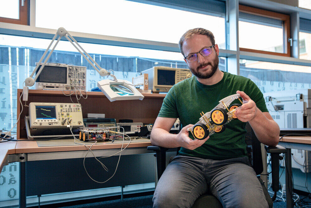 Ein Student hält ein Roboterfahrgestell in der Hand und schaut in die Kamera.
