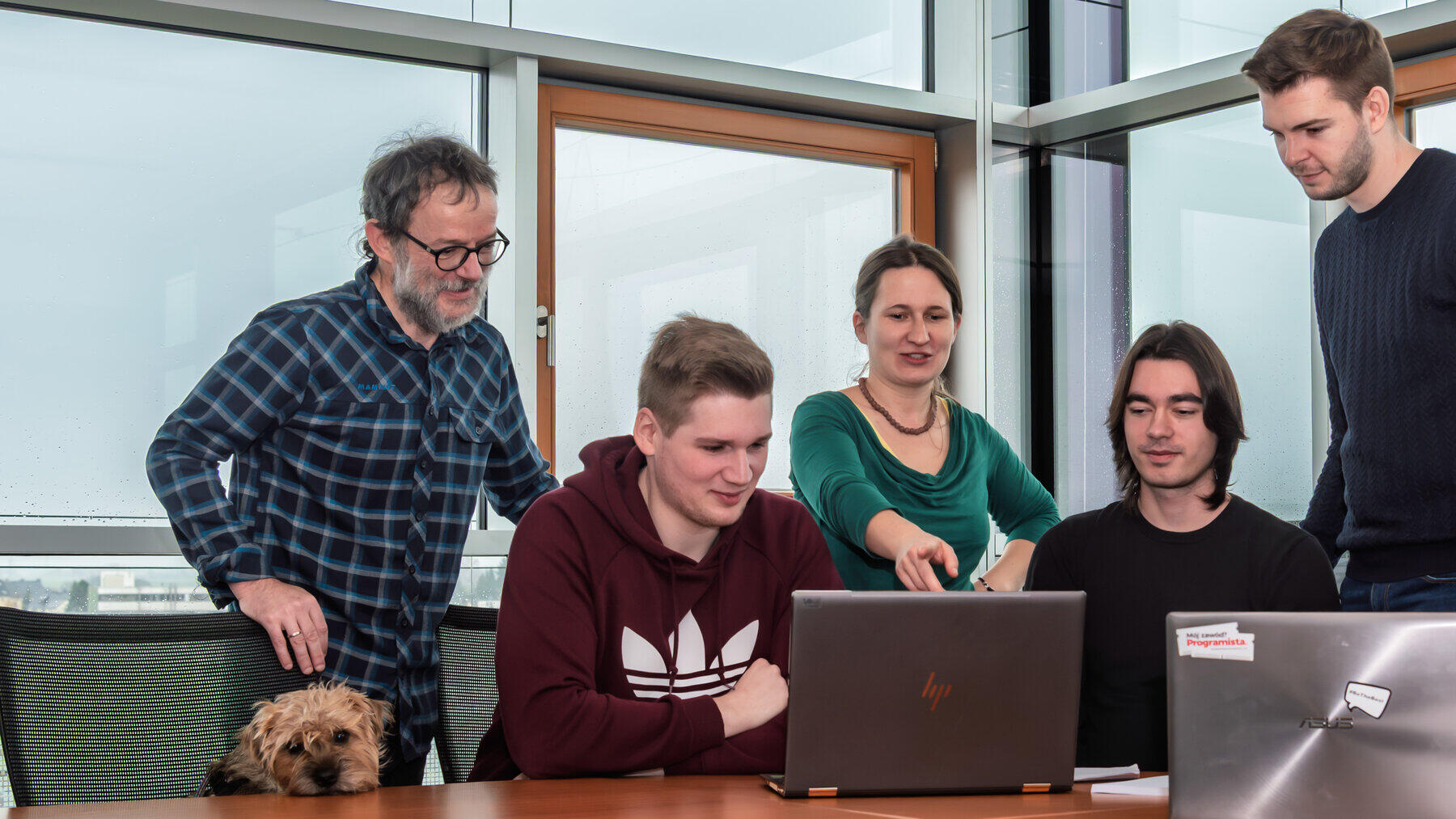 Vier Personen stehen und sitzen an einem Tisch mit einem Laptop