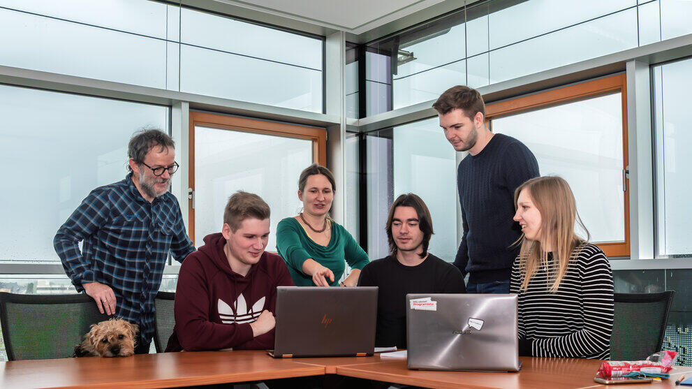 Vier Personen stehen und sitzen an einem Tisch mit einem Laptop