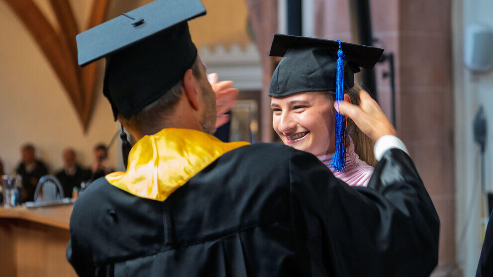 A professor in a black gown places a hat on a smiling graduate’s head.