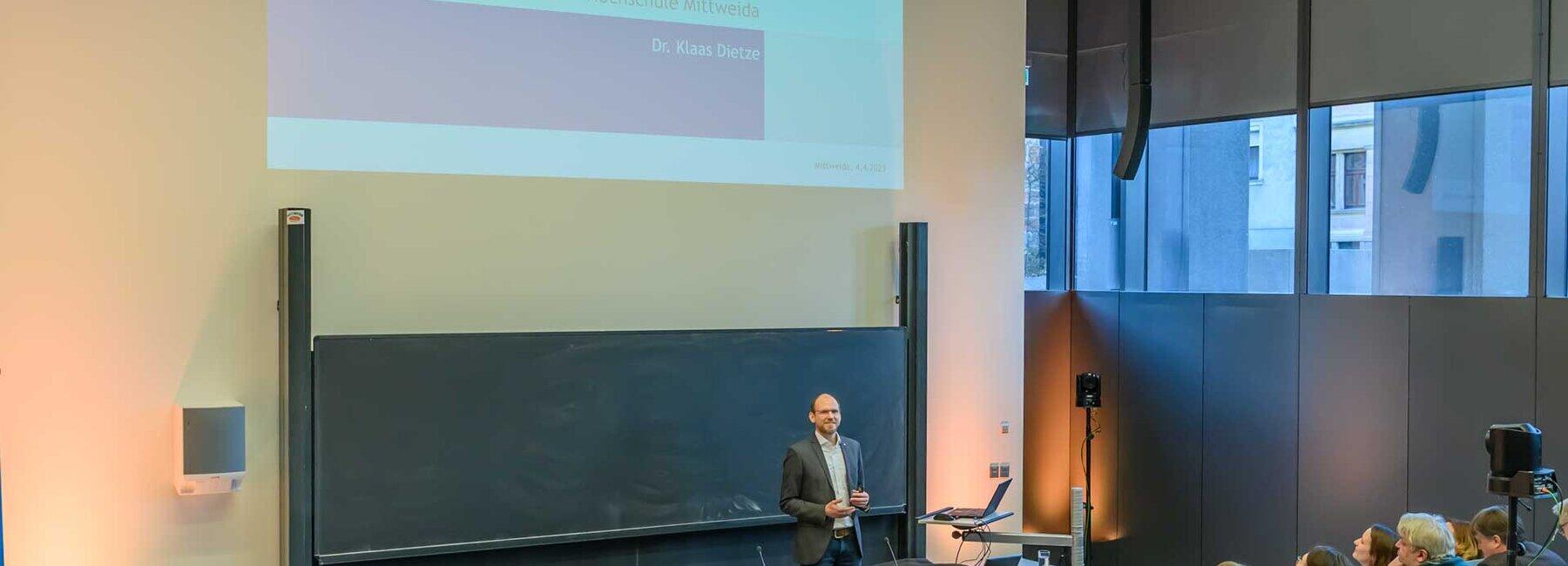 : Blick von seitlich hinten über die Köpfe der Zuschauenden auf den Dozenten, der vor der Tafel steht. Über ihm zeigt die Projektion die Titelfolie der Vorlesung: „One Health – Zeitenwende, wie wir Gesundheit verstehen? 5. Öffentliche Ringvorlesung. Hochschule Mittweida. Dr. Klaas Dietze.“