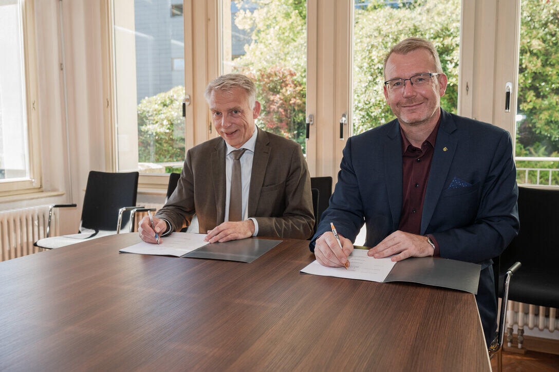 Zwei männliche Personen im Anzug sitzen nebeneinander an einem Tisch und blicken nach vollzogener Unterschrift auf den Papieren vor ihnen freundlich in die Kamera.