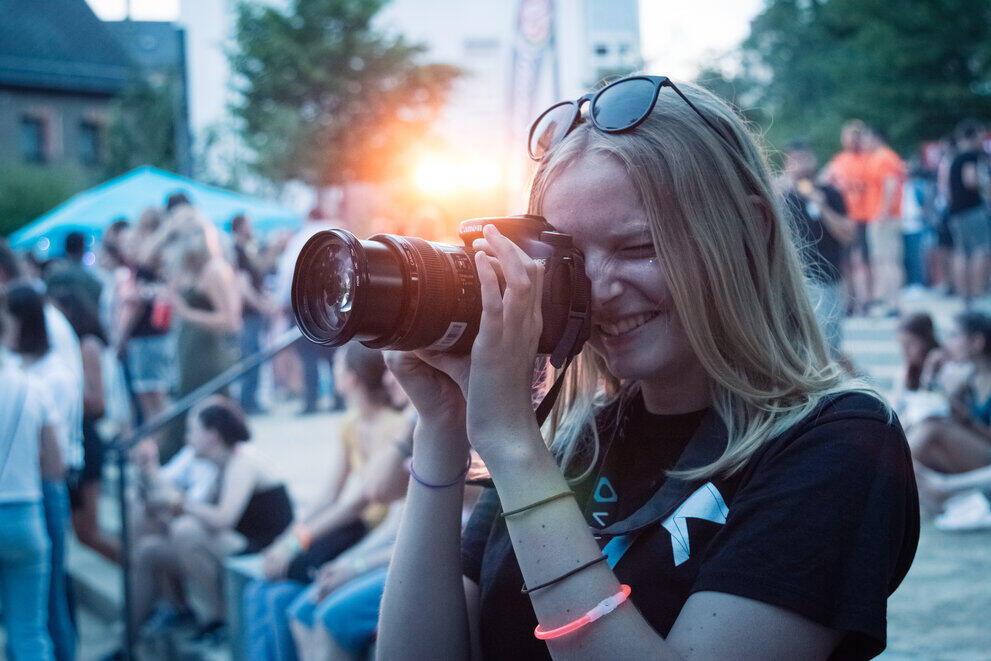 Eine Studentin fotografiert mit einer Kamera, während im Hintergrund viele Menschen sitzen und die Sonne untergeht.