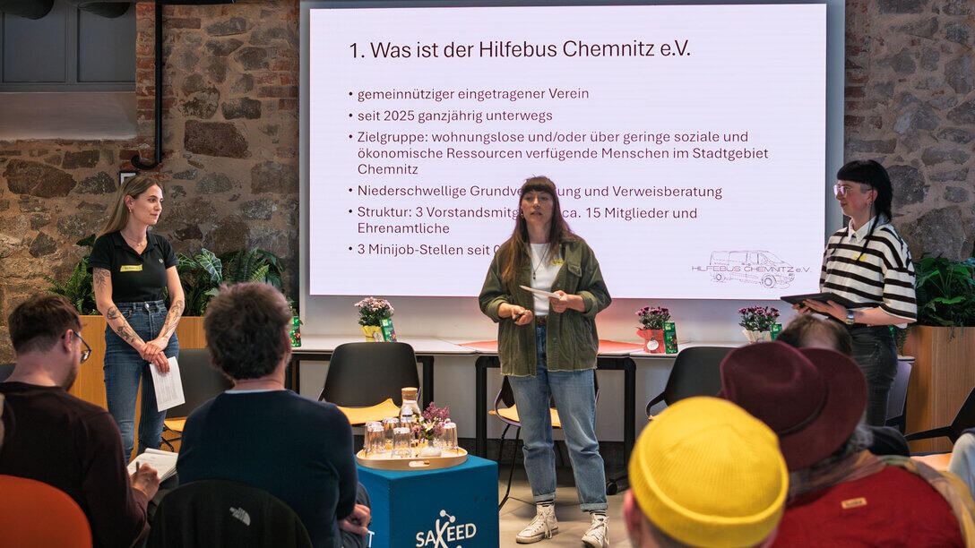Drei weibliche Personen stehen vor einem Screen. Die mittlere Person spricht zum Publikum. Auf dem Screen ist eine Folie mit der Überschrift „Was ist der Hilfebus Chemnitz e.V.“ und weiteren Anstrichen zu sehen.
