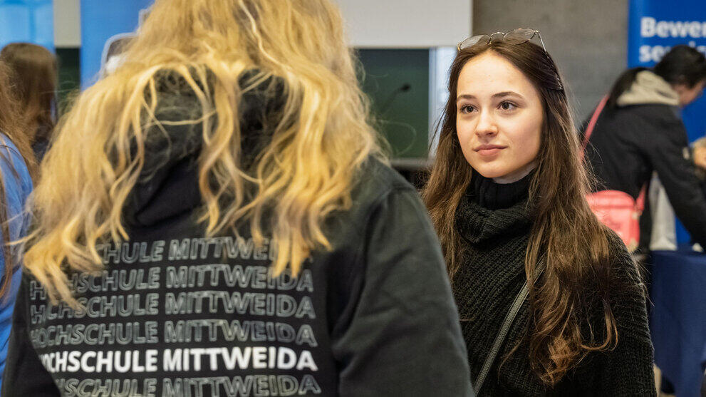 Two women are talking. The words ‘Mittweida University of Applied Sciences’ can be seen several times on one of their hoodies.