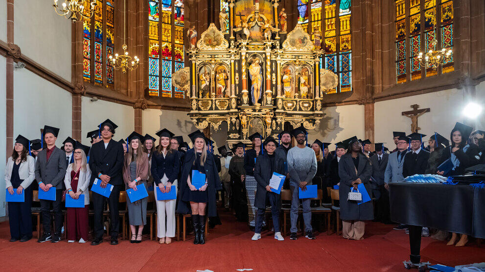 Graduates wearing caps and carrying blue certificate folders are standing in the chancel.