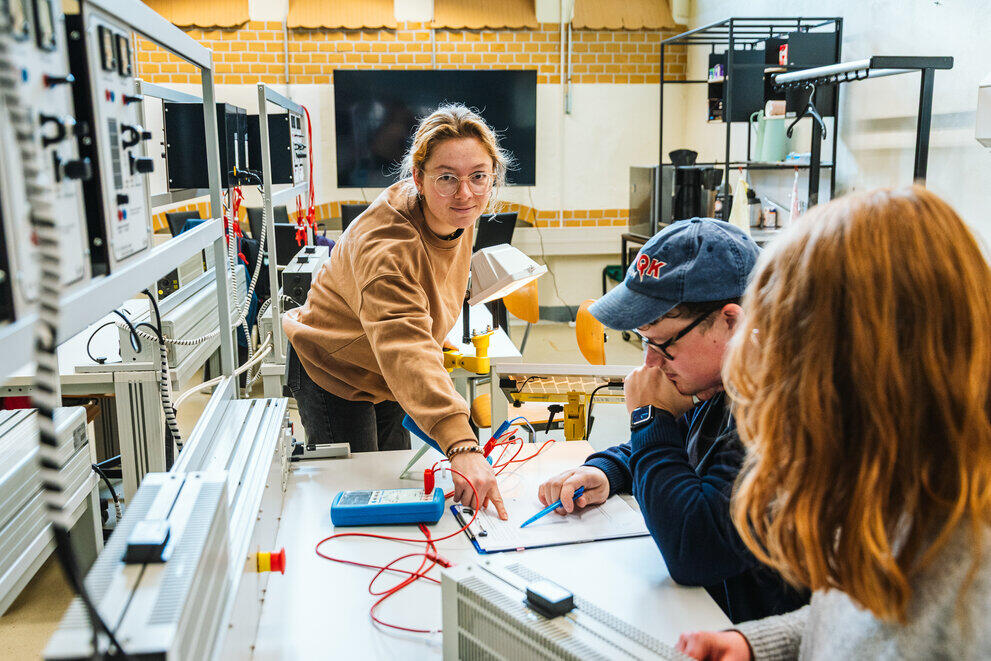 Drei Studierende sitzen und stehen in einem Elektrotechnik-Labor und zeigen auf ein Klemmbrett mit Messdaten.