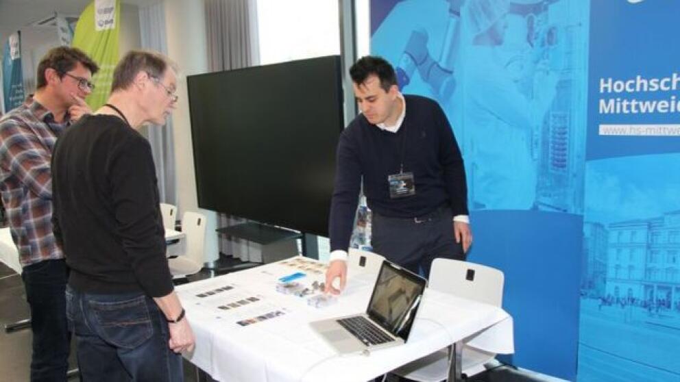 Messestand, zwei männliche Personen lassen sich von einer männlichen Person etwas zeigen