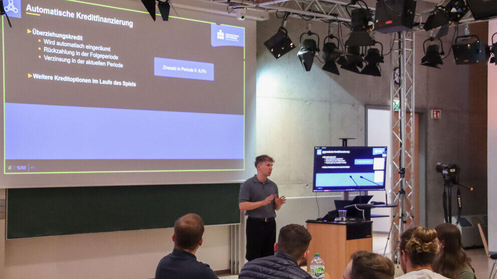 A young man is standing between a screen and a projection screen, both of which are displaying the same content: a slide with the heading ‘Automatic Credit Financing’.