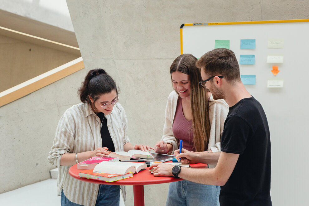Three students are standing at a table covered in books and studying together