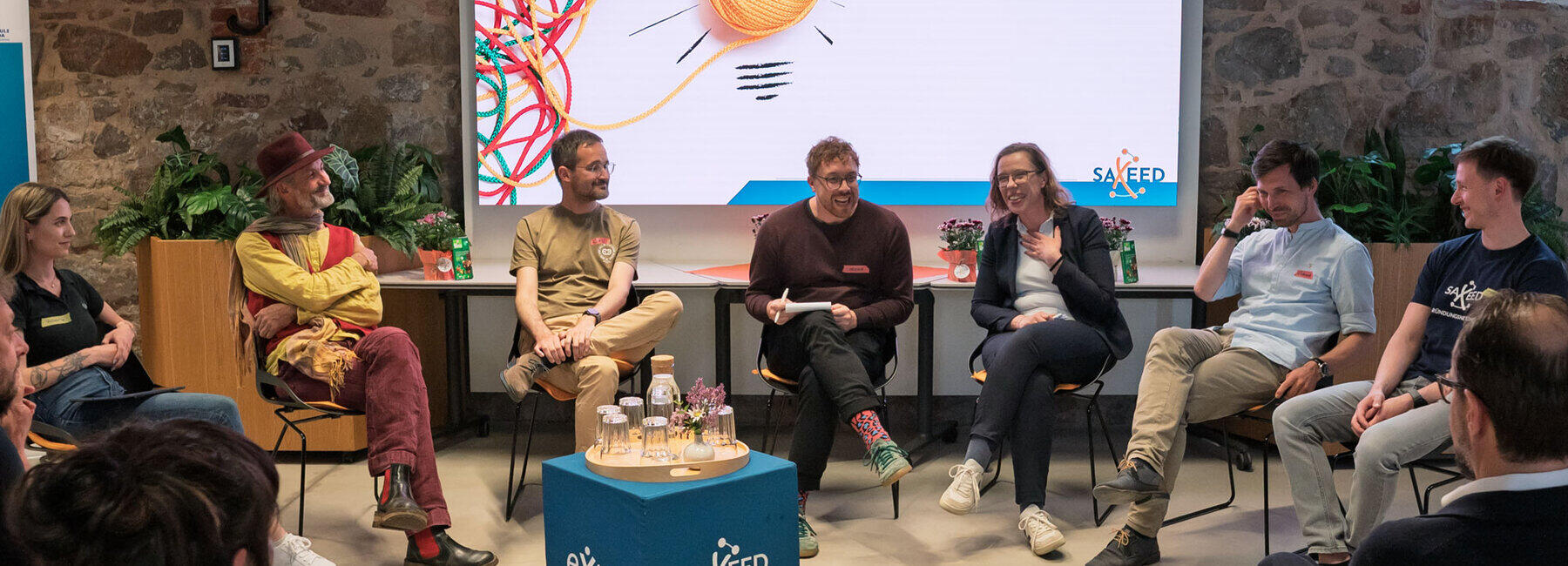 Seven people sitting on chairs in front of a large screen displaying the words ‘Social Entrepreneurship: How to turn social impact into a viable business model’. In the foreground of the picture, the heads of the audience members can be seen from behind.