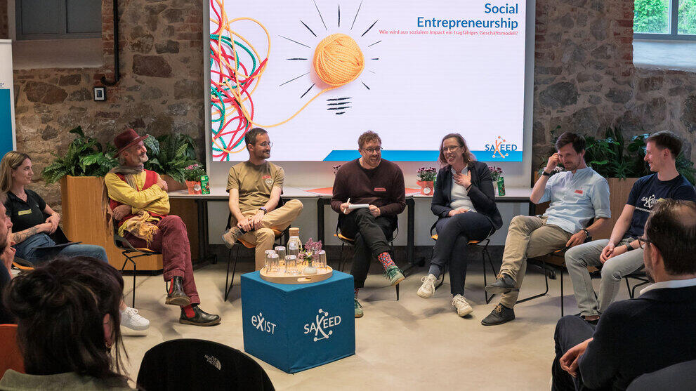 Sieben Personen auf Stühlen vor einem großen Screen, auf dem „Social Entrepreneurship. Wir wird aus sozialem Impact ein tragfähiges Geschäftsmodell“ zu lesen ist. Im Bildvordergrund sind Köpfe von Zuschauenden von hinten zu sehen.
