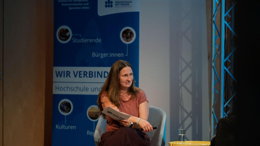 Weibliche Person im Sessel mit Moderationskarten. Im Hintergrund steht ein blaues Rollup mit unter anderem dem Text „Wir verbinden“.