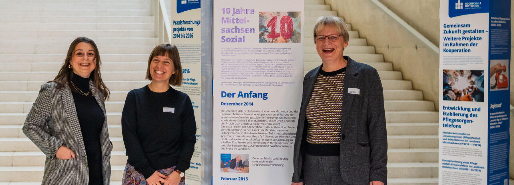 Three women laughing happily. Behind them are rectangular display stands, one of which bears the inscription ‘10 Years of Mittelsachsen Sozial’ and further text.